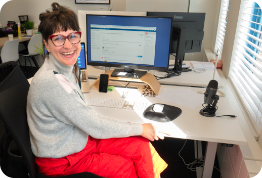 Woman smiling at her desk with a computer, microphone, and papers in a bright office.