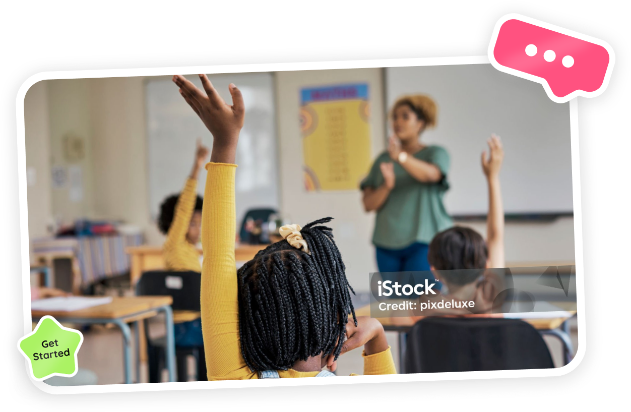 Children raising hands in a classroom while a teacher stands at the front, blurred in the background.