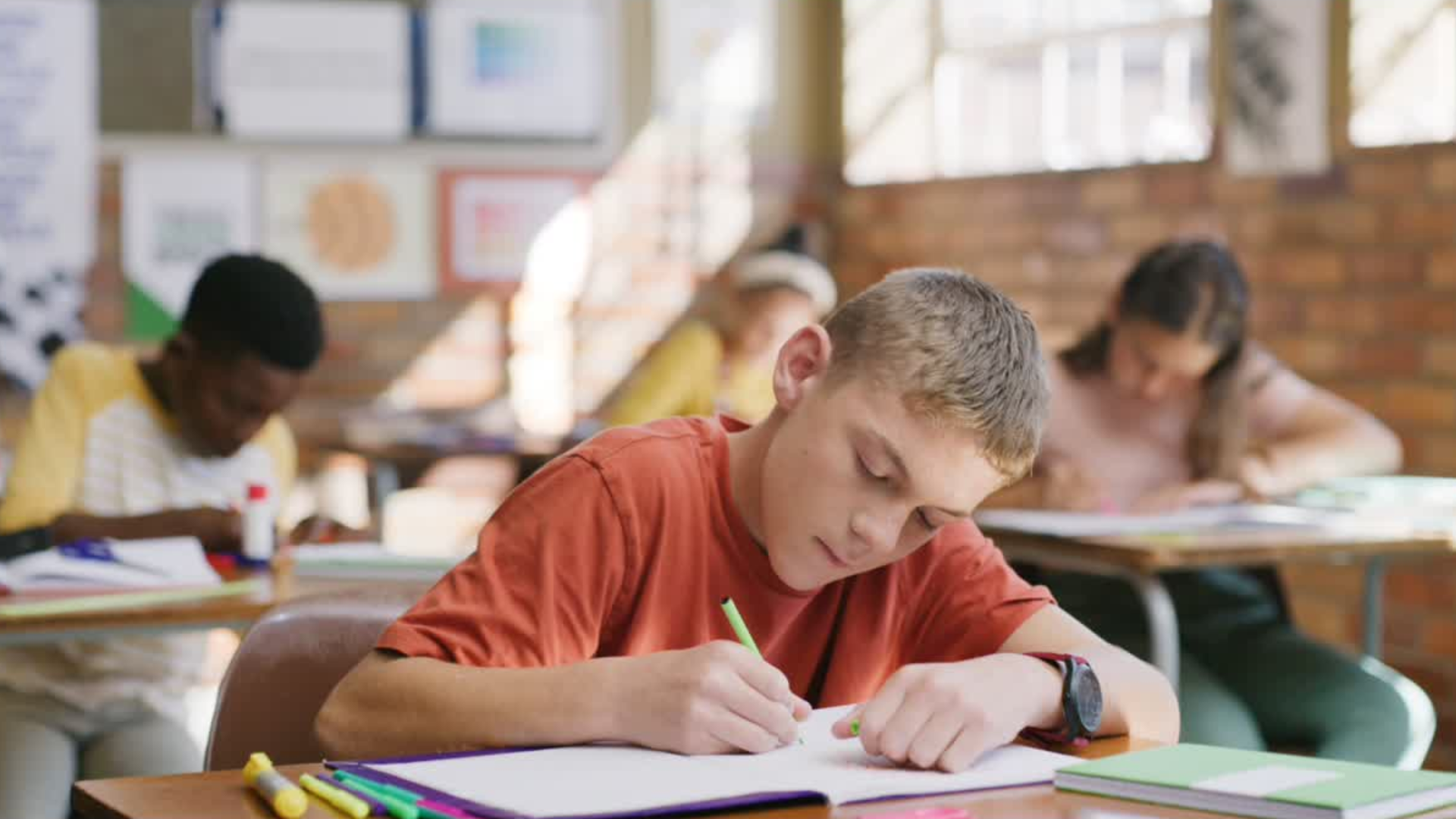A boy in a red shirt writes in a notebook at his desk in a classroom with other students.